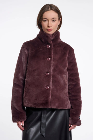 A woman wearing a plush, cropped faux fur jacket in a deep plum-brown color with matching buttons, paired with a black skirt, against a plain white studio background.