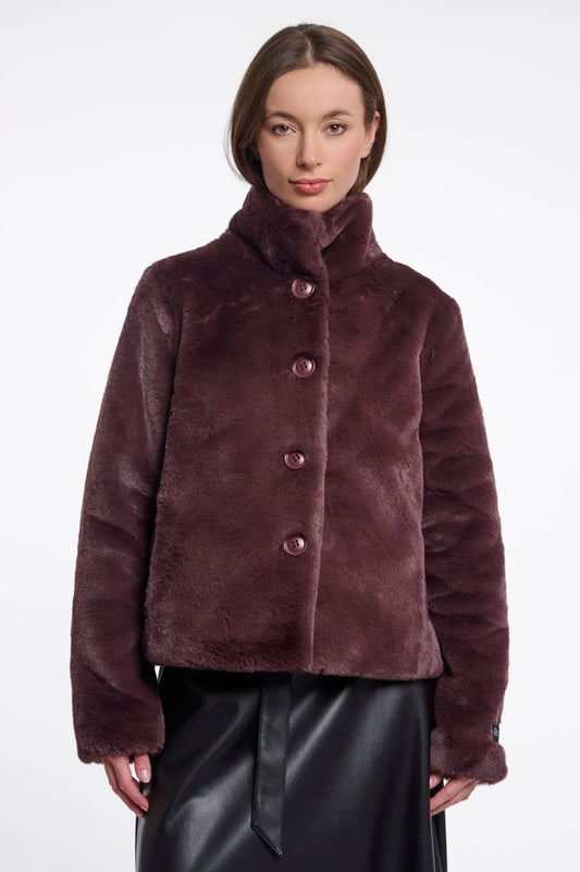 A woman wearing a plush, cropped faux fur jacket in a deep plum-brown color with matching buttons, paired with a black skirt, against a plain white studio background.