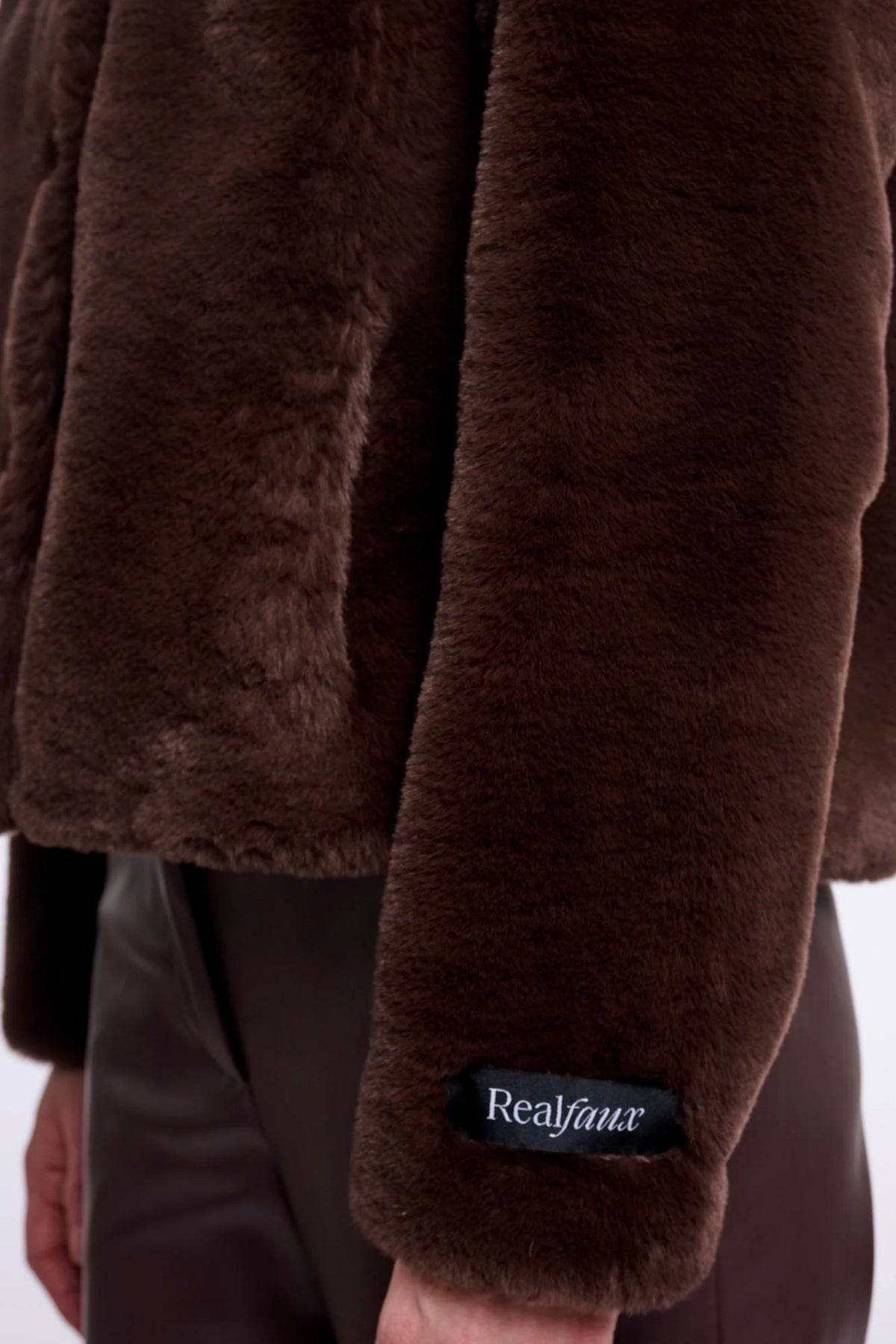 Close-up of a brown faux fur jacket with a black brand label reading 'Realfaux' on the cuff, worn with dark brown pants.