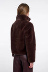 A model shown from the back wearing a dark brown plush faux fur jacket and dark brown pants against a plain white studio background.