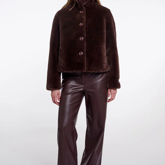 A model wearing a dark brown cropped faux-fur jacket with a high collar and three front buttons, paired with dark brown faux-leather pants and black pointed shoes, against a light gray studio background.
