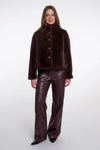 A model wearing a dark brown cropped faux-fur jacket with a high collar and three front buttons, paired with dark brown faux-leather pants and black pointed shoes, against a light gray studio background.