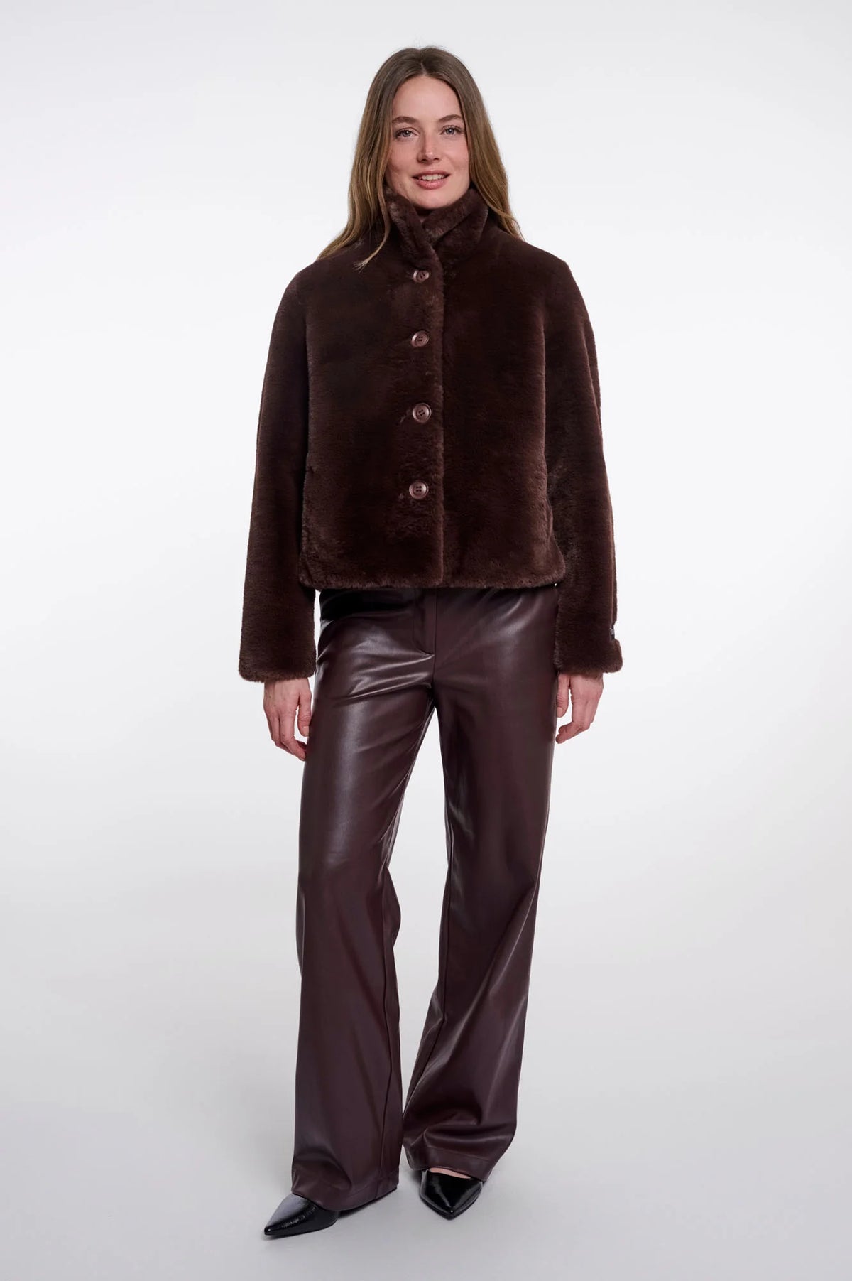 A model wearing a dark brown cropped faux-fur jacket with a high collar and three front buttons, paired with dark brown faux-leather pants and black pointed shoes, against a light gray studio background.
