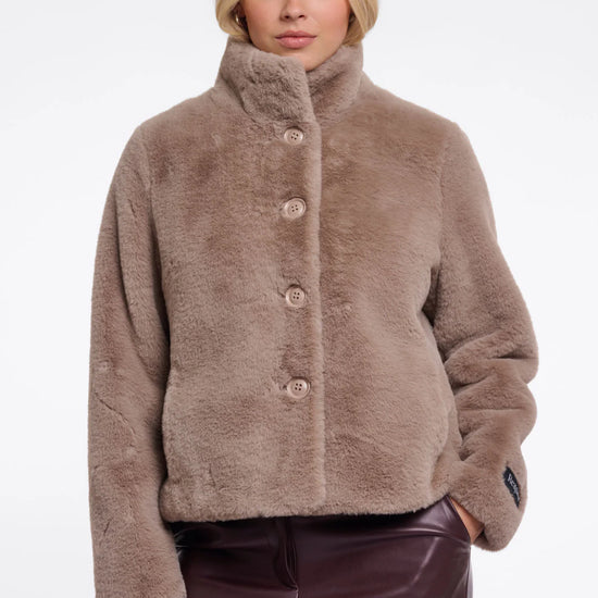 A woman modeling a cropped, high-neck faux fur jacket in a warm taupe-brown color, paired with dark brown faux-leather pants, against a plain white studio background.