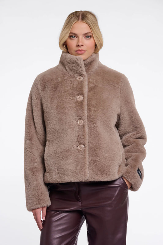 A woman modeling a cropped, high-neck faux fur jacket in a warm taupe-brown color, paired with dark brown faux-leather pants, against a plain white studio background.