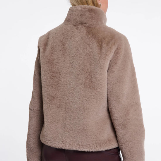 A woman viewed from the back wearing a soft, fuzzy cropped jacket in a taupe-brown color, paired with dark brown pants, against a plain white studio background.
