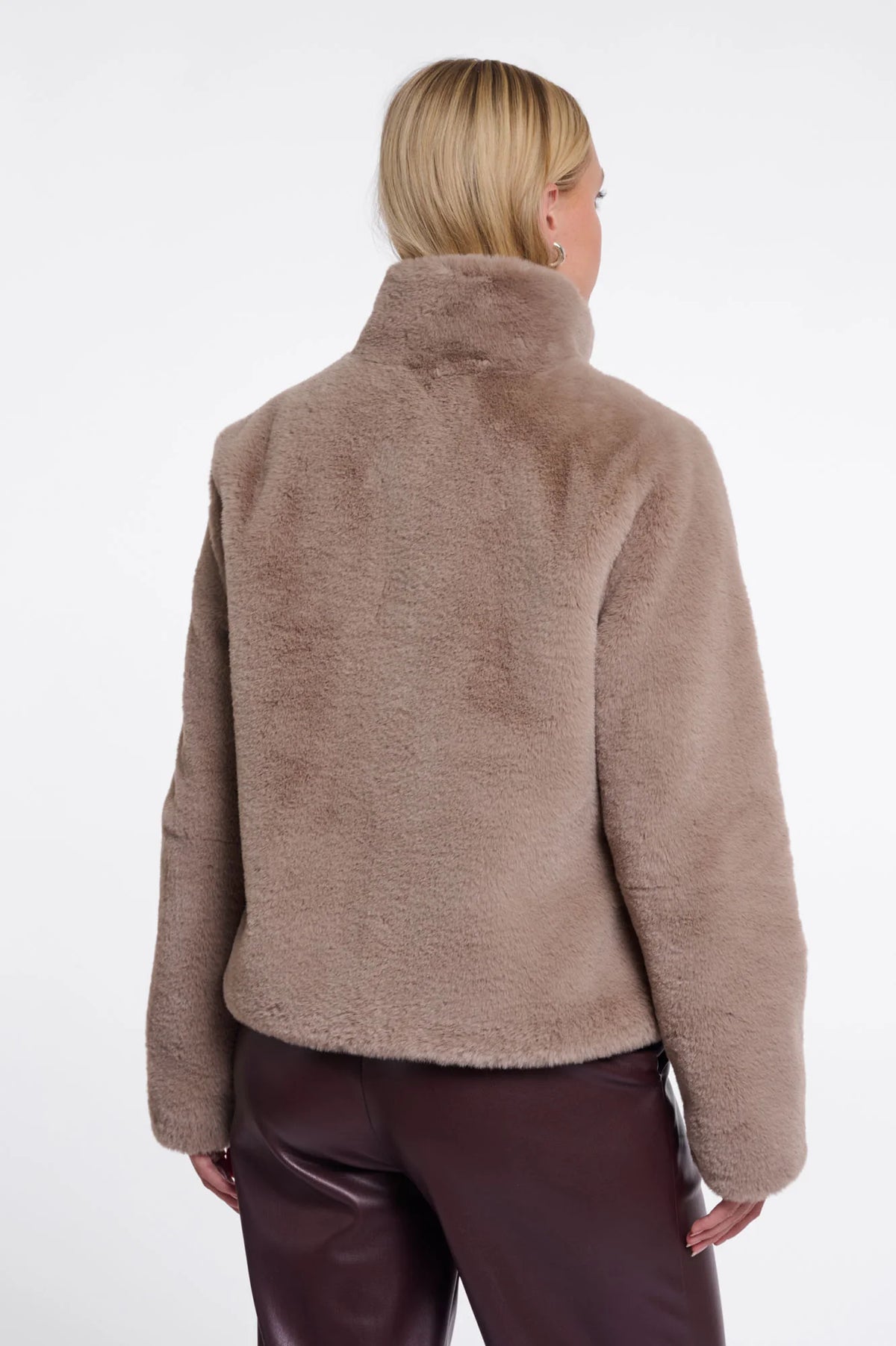 A woman viewed from the back wearing a soft, fuzzy cropped jacket in a taupe-brown color, paired with dark brown pants, against a plain white studio background.