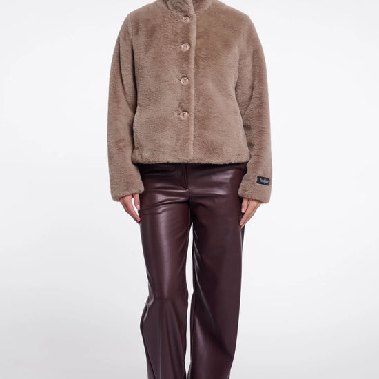 A model stands against a light gray studio background wearing a taupe-brown fuzzy button-front jacket with a high stand collar and dark brown faux-leather pants.