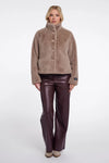 A model stands against a light gray studio background wearing a taupe-brown fuzzy button-front jacket with a high stand collar and dark brown faux-leather pants.
