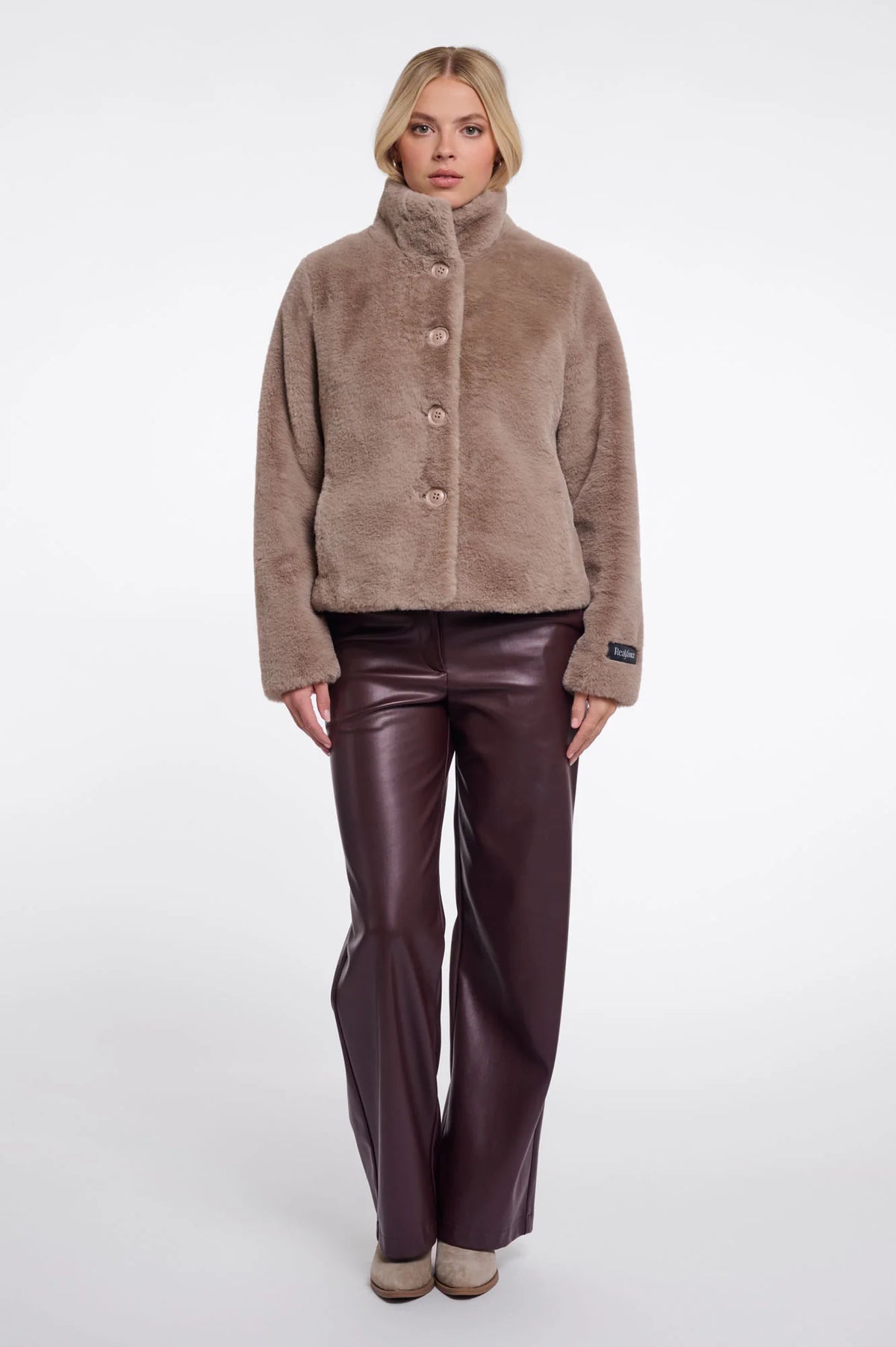 A model stands against a light gray studio background wearing a taupe-brown fuzzy button-front jacket with a high stand collar and dark brown faux-leather pants.