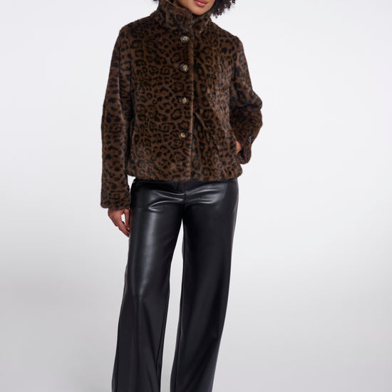 A model wearing a brown and black leopard-print faux fur jacket with a high collar, paired with black faux leather wide-leg pants and black pointed-toe heels, photographed against a light gray studio background.