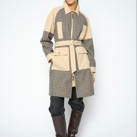 A model wearing a patchwork coat with beige and brown gingham patterns, paired with dark pants and brown leather boots, against a light gray studio background.