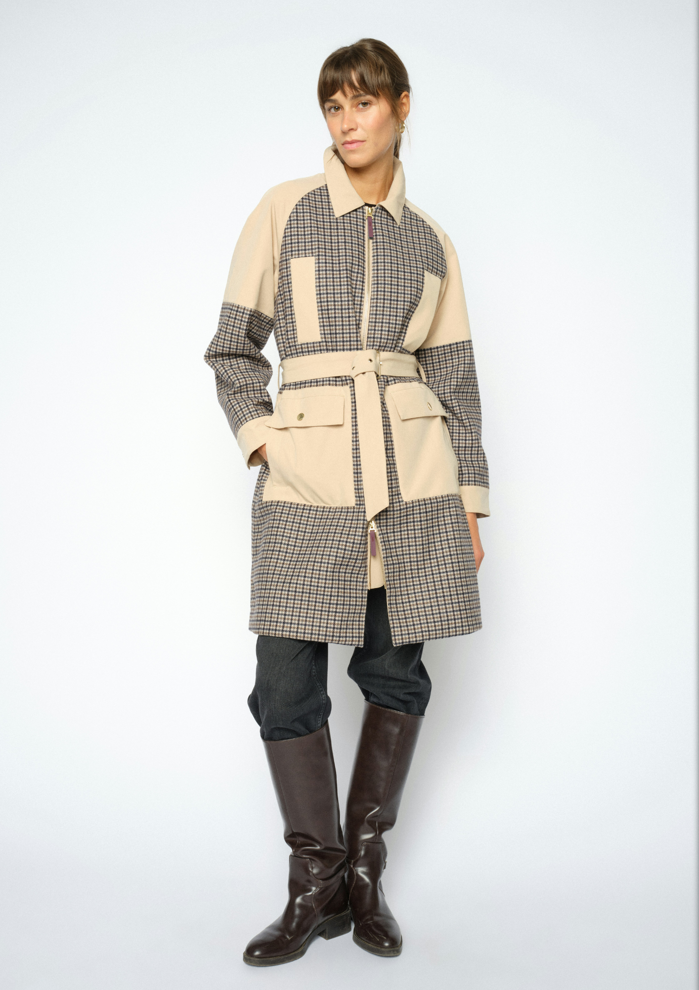 A model wearing a patchwork coat with beige and brown gingham patterns, paired with dark pants and brown leather boots, against a light gray studio background.