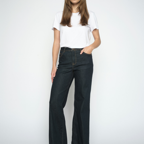 A woman modeling a pair of dark-wash wide-leg jeans with a white t-shirt and brown suede boots, standing against a light gray studio background.