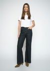 A woman modeling a pair of dark-wash wide-leg jeans with a white t-shirt and brown suede boots, standing against a light gray studio background.