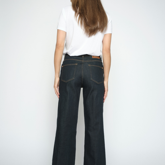 A woman viewed from the back wearing dark wash flare jeans, a white t-shirt, and heels against a light gray studio background.