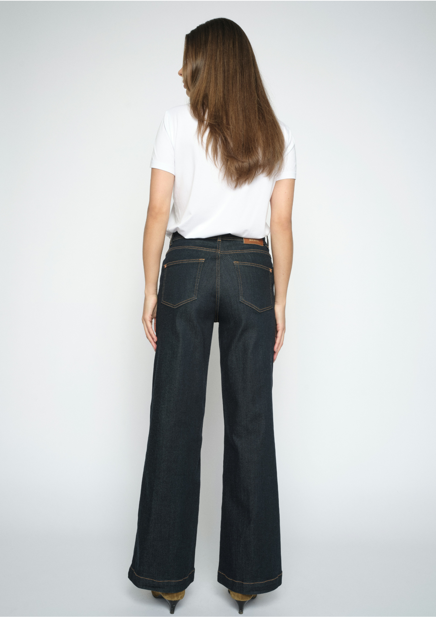 A woman viewed from the back wearing dark wash flare jeans, a white t-shirt, and heels against a light gray studio background.