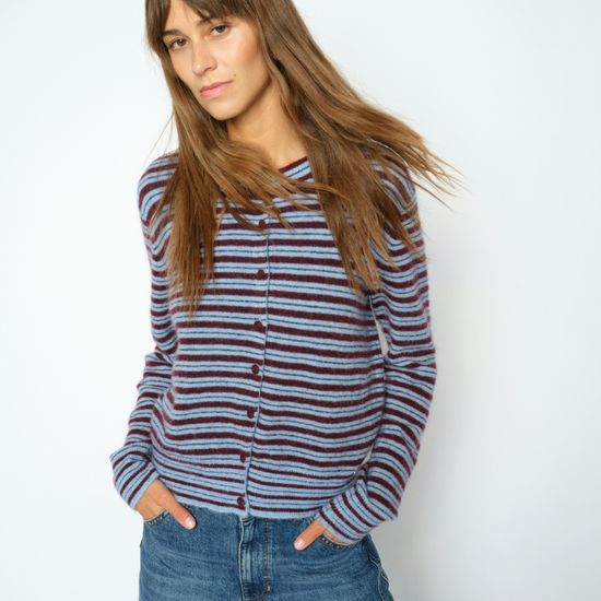 A woman with long brown hair wearing a striped button-up cardigan with blue jeans, standing against a plain light gray background.