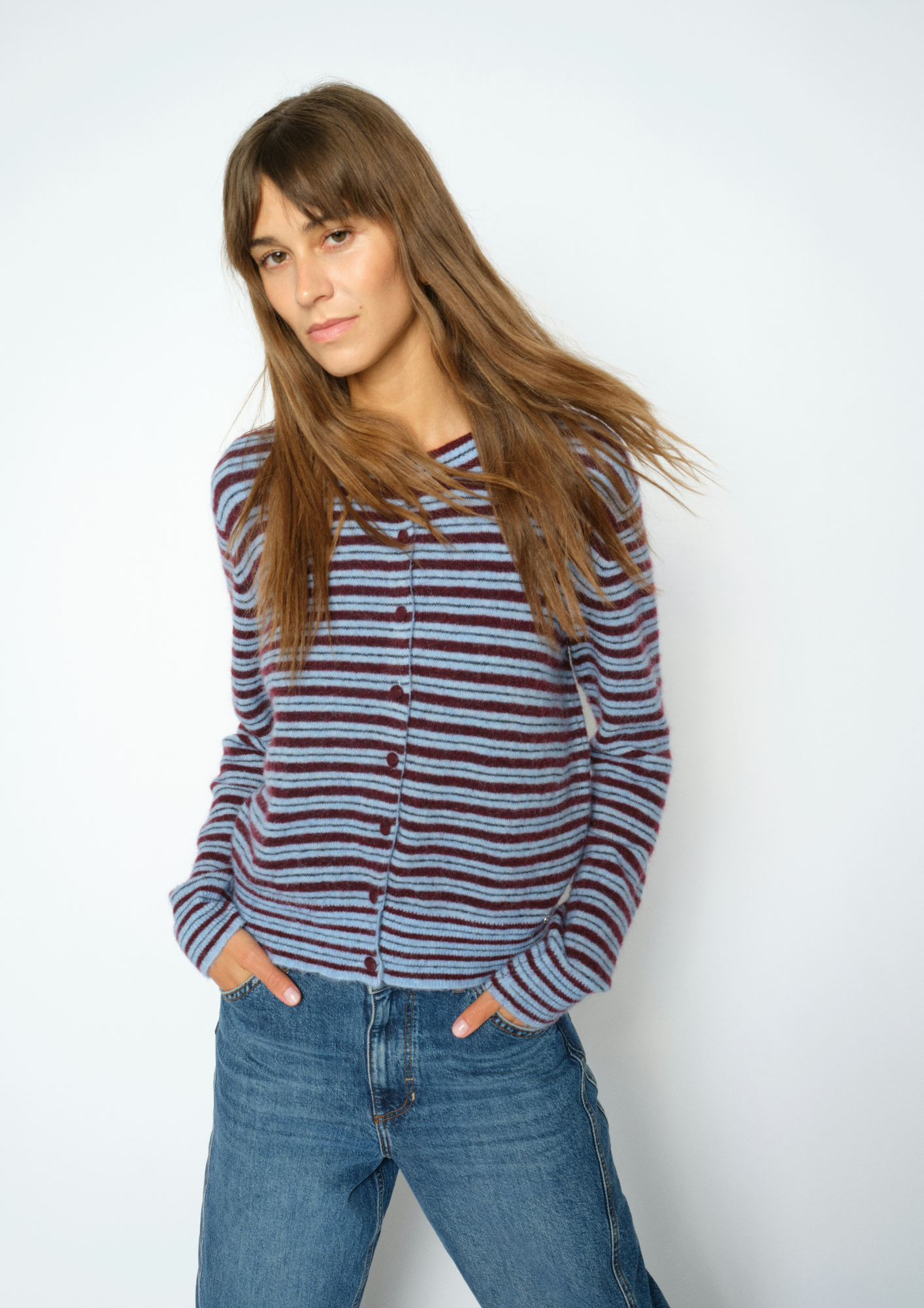 A woman with long brown hair wearing a striped button-up cardigan with blue jeans, standing against a plain light gray background.