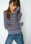 A woman wearing a fuzzy knit cardigan with horizontal stripes in dusty blue, burgundy, and cream, paired with blue jeans, posing against a plain white wall.