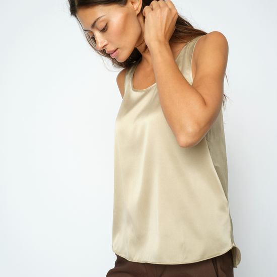 A woman wearing a sleeveless beige satin top and dark brown pants, posing against a light gray studio background.
