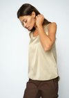 A woman wearing a sleeveless beige satin top and dark brown pants, posing against a light gray studio background.