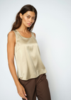 A woman modeling a sleeveless satin top in a light champagne beige color, paired with brown trousers, against a plain light gray studio background.