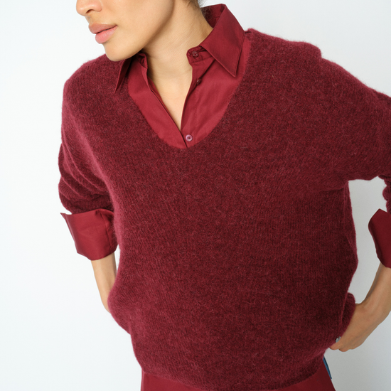 A woman wearing a burgundy knit v-neck sweater layered over a matching burgundy collared button-up shirt, paired with blue jeans, against a plain white background.