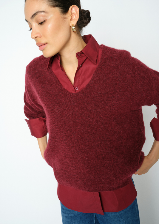 A woman wearing a burgundy knit v-neck sweater layered over a matching burgundy collared button-up shirt, paired with blue jeans, against a plain white background.