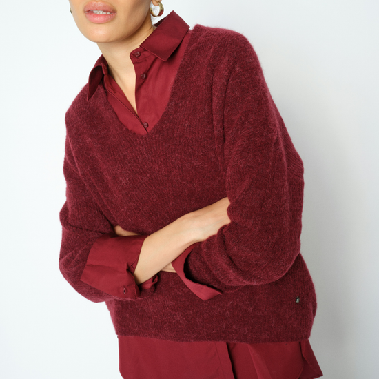 A woman wearing a textured deep burgundy knit sweater over a matching burgundy collared blouse and blue jeans, photographed against a plain light gray studio background.