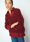 A woman wearing a textured deep burgundy knit sweater over a matching burgundy collared blouse and blue jeans, photographed against a plain light gray studio background.