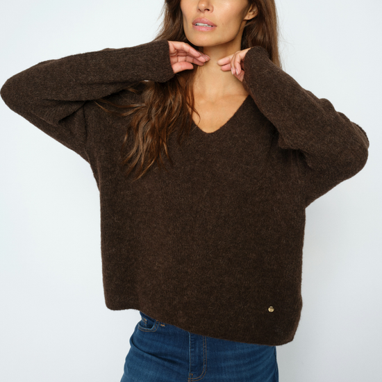 A woman wearing a dark brown fuzzy knit sweater with a V-neck and wide sleeves, paired with blue jeans, posing against a light gray studio background.