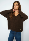 A woman wearing a dark brown fuzzy knit sweater with a V-neck and wide sleeves, paired with blue jeans, posing against a light gray studio background.