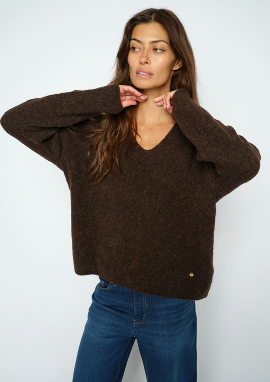 A woman wearing a dark brown fuzzy knit sweater with a V-neck and wide sleeves, paired with blue jeans, posing against a light gray studio background.