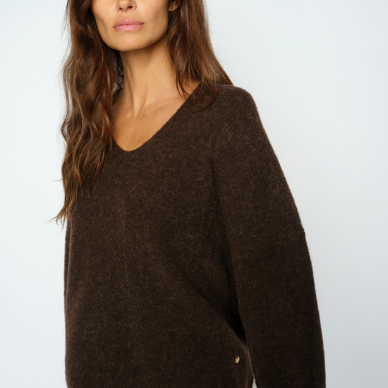 A woman wearing a dark brown fuzzy knit sweater with a wide V-neck and long sleeves, paired with blue jeans, photographed against a light gray studio background.