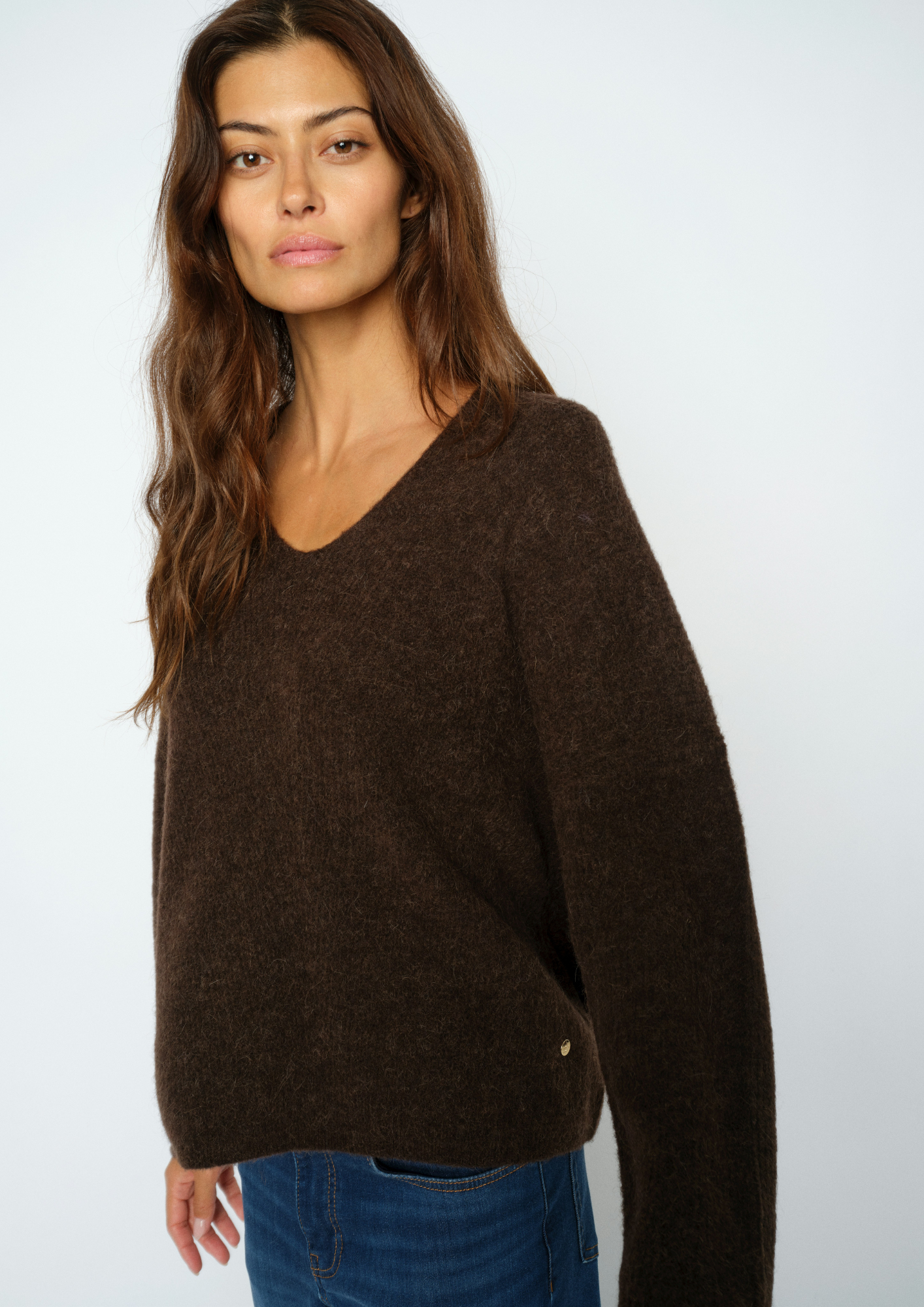 A woman wearing a dark brown fuzzy knit sweater with a wide V-neck and long sleeves, paired with blue jeans, photographed against a light gray studio background.