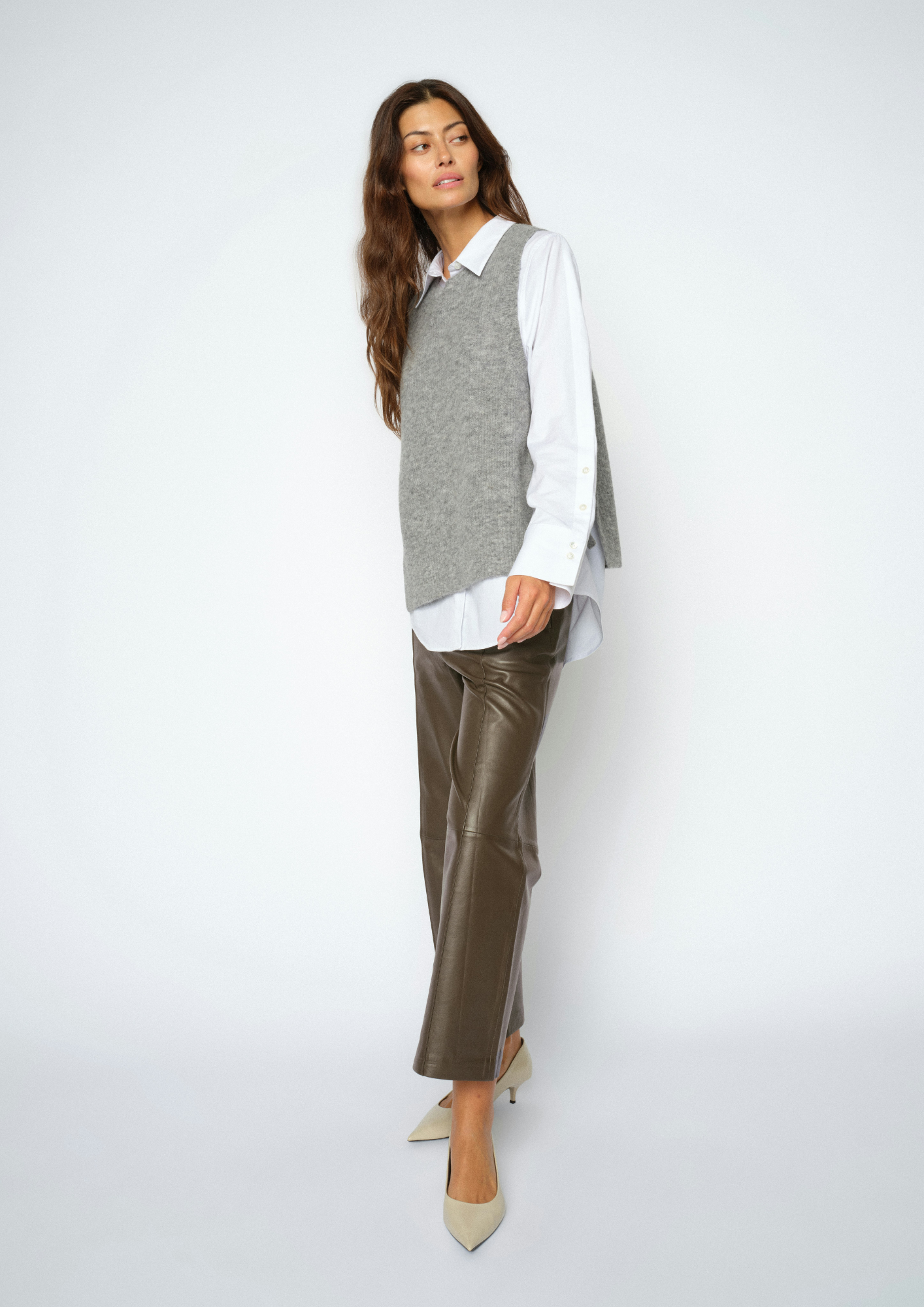 A woman modeling a gray knit sweater vest over a white collared shirt, paired with brown leather cropped pants and beige pointed-toe heels, against a light gray studio background.