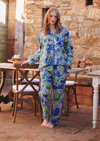 A woman stands barefoot in a rustic outdoor patio setting wearing a blue pajama set with a green and white paisley print. The background features a stone wall, wooden dining tables, and chairs.