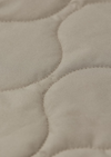 Close-up of a quilted fabric surface in a warm light beige or oatmeal tone, showing stitched oval sections and soft texture.