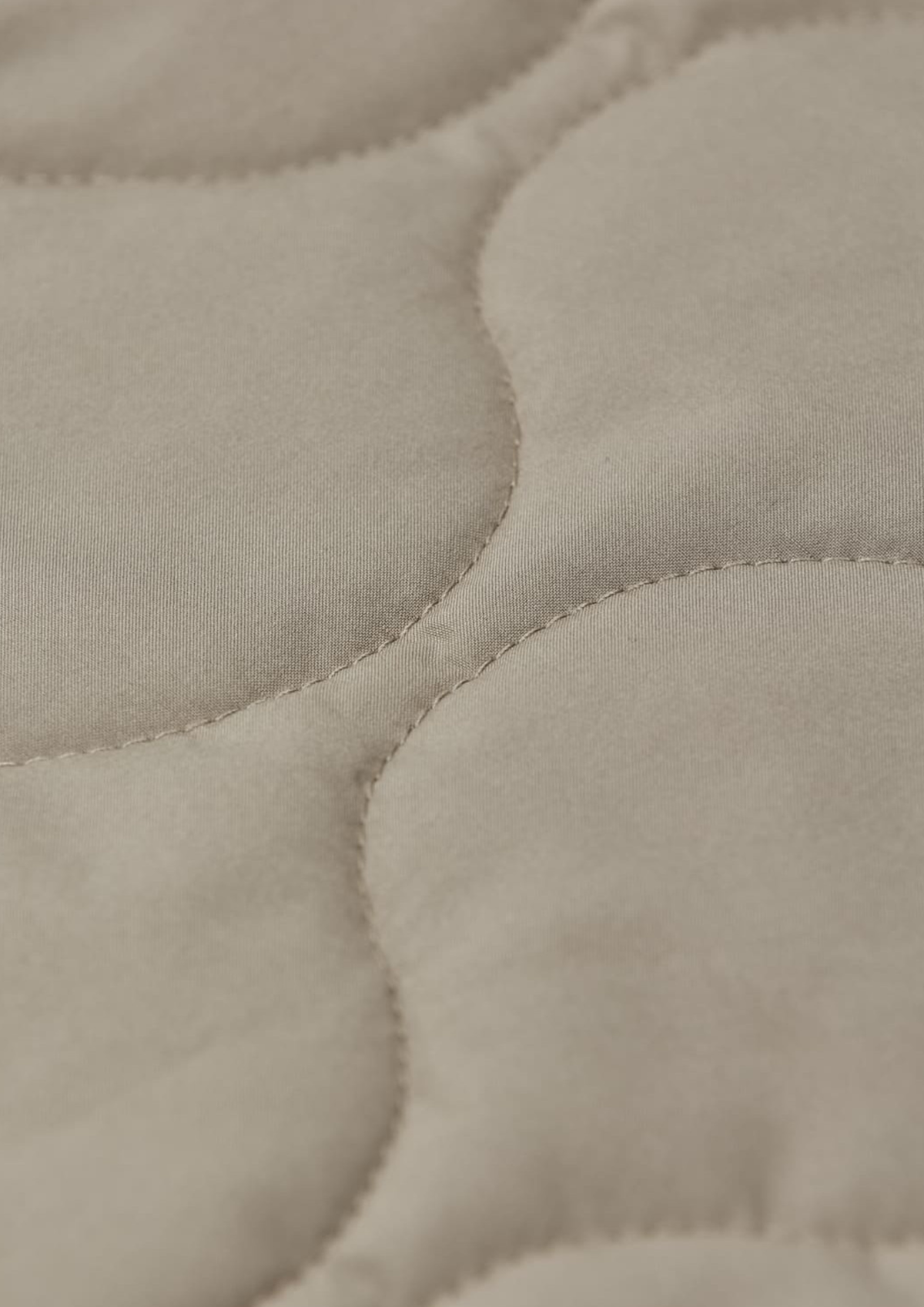 Close-up of a quilted fabric surface in a warm light beige or oatmeal tone, showing stitched oval sections and soft texture.