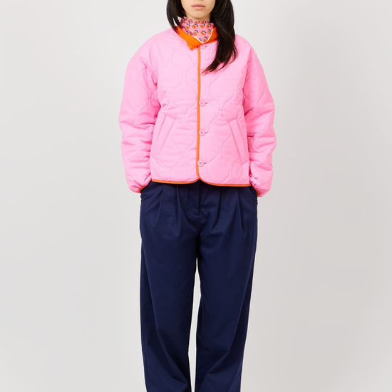 A model stands against a light gray studio background wearing a bright pink quilted jacket with orange trim, navy wide-leg pants, and patterned sneakers.