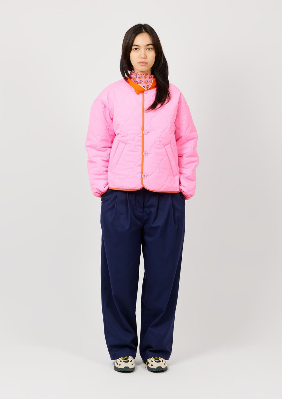 A model stands against a light gray studio background wearing a bright pink quilted jacket with orange trim, navy wide-leg pants, and patterned sneakers.