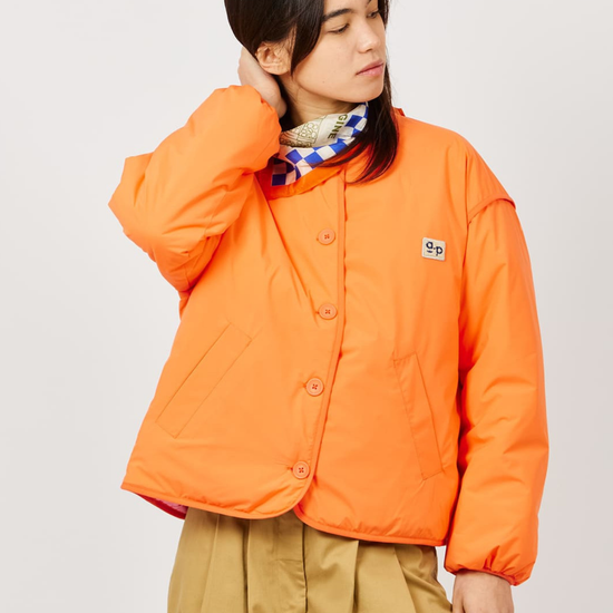 A model wearing a bright orange cropped puffer jacket with a small 'Sp' logo patch on the chest, styled with tan pants and a patterned scarf, against a light gray studio background.
