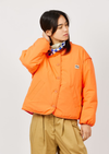 A model wearing a bright orange cropped puffer jacket with a small 'Sp' logo patch on the chest, styled with tan pants and a patterned scarf, against a light gray studio background.