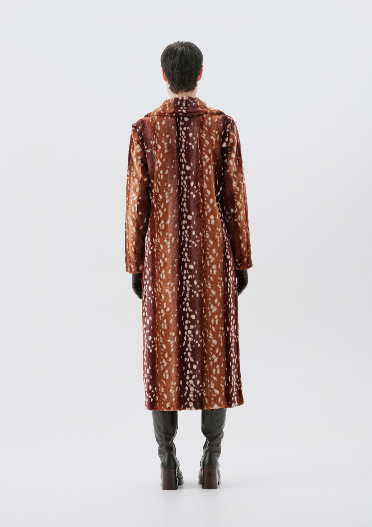 A model viewed from the back wearing a long brown coat with a white floral pattern, paired with dark gray pants and brown boots, set against a plain light gray studio background.