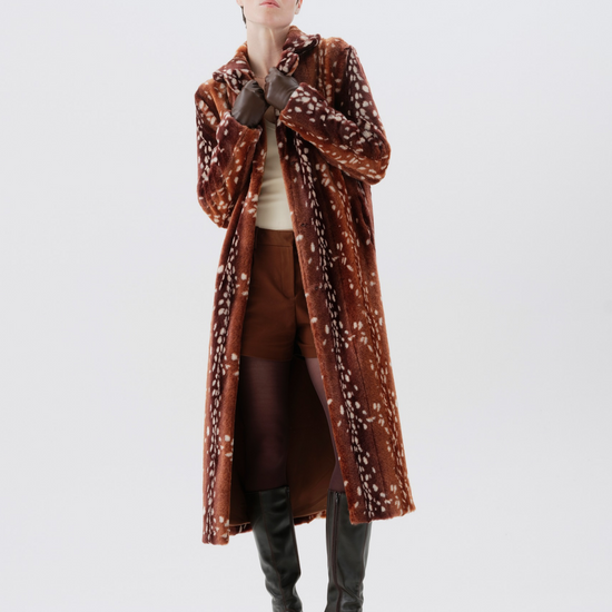 A model wearing a long brown velvet coat with a white floral pattern, styled with brown trousers, a cream top, and black knee-high boots against a plain white studio background.