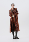 A model wearing a long brown velvet coat with a white floral pattern, styled with brown trousers, a cream top, and black knee-high boots against a plain white studio background.