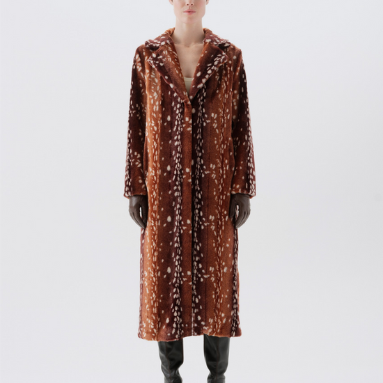 A model stands against a light gray studio background wearing a long brown coat with a white abstract pattern, black gloves, and black boots.