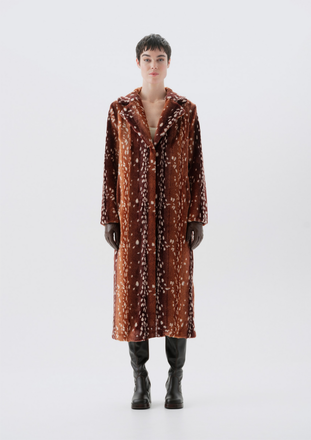 A model stands against a light gray studio background wearing a long brown coat with a white abstract pattern, black gloves, and black boots.