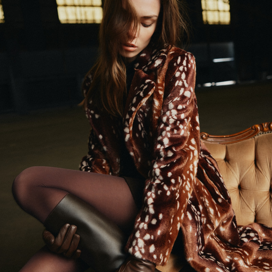 A woman sits on a tufted sofa in a dimly lit interior, wearing a brown faux-fur coat with a white spotted pattern, dark tights, and tall dark boots.